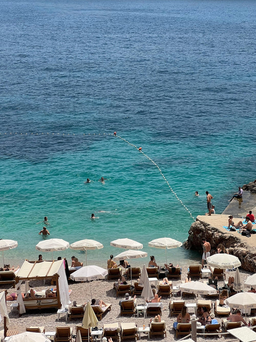 Beach scene with people, umbrellas, and chairs near a rocky outcrop with clear blue water.