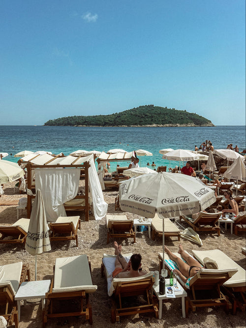 Beach scene with people under Coca-Cola umbrellas and chairs by the ocean.