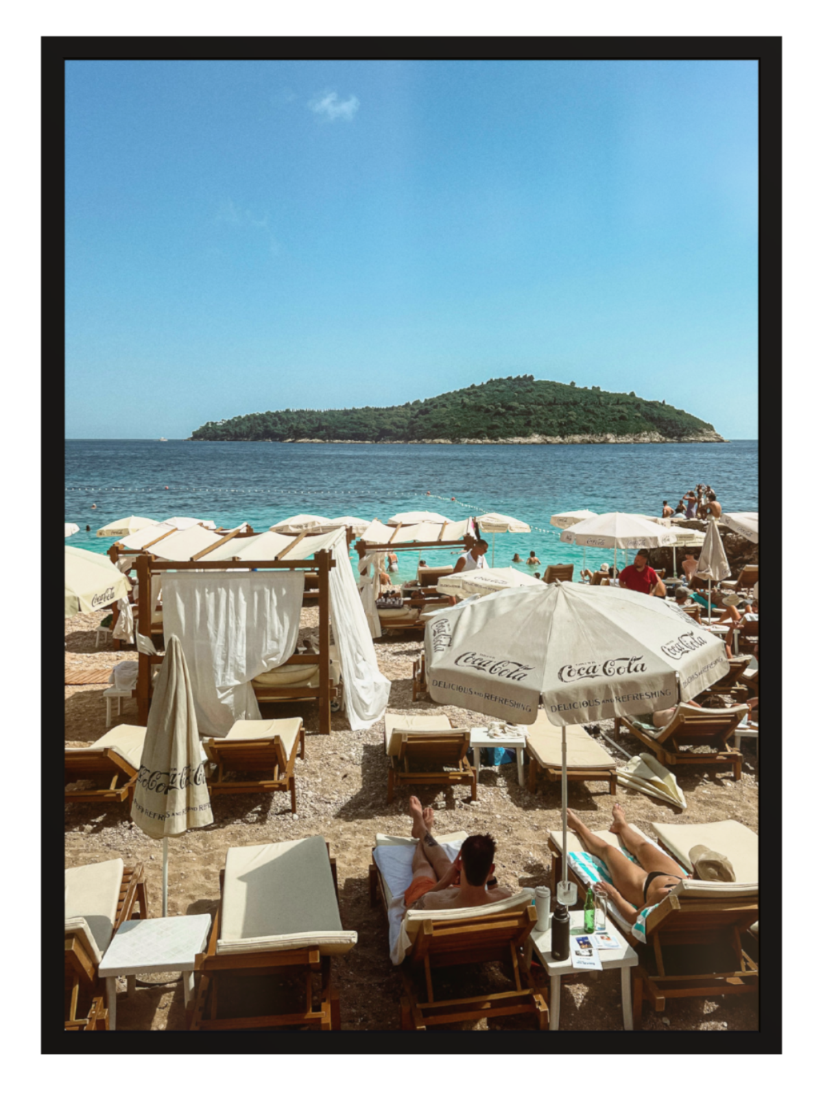 Beach scene with people under umbrellas and chairs, Coca-Cola umbrella visible, clear blue sky.