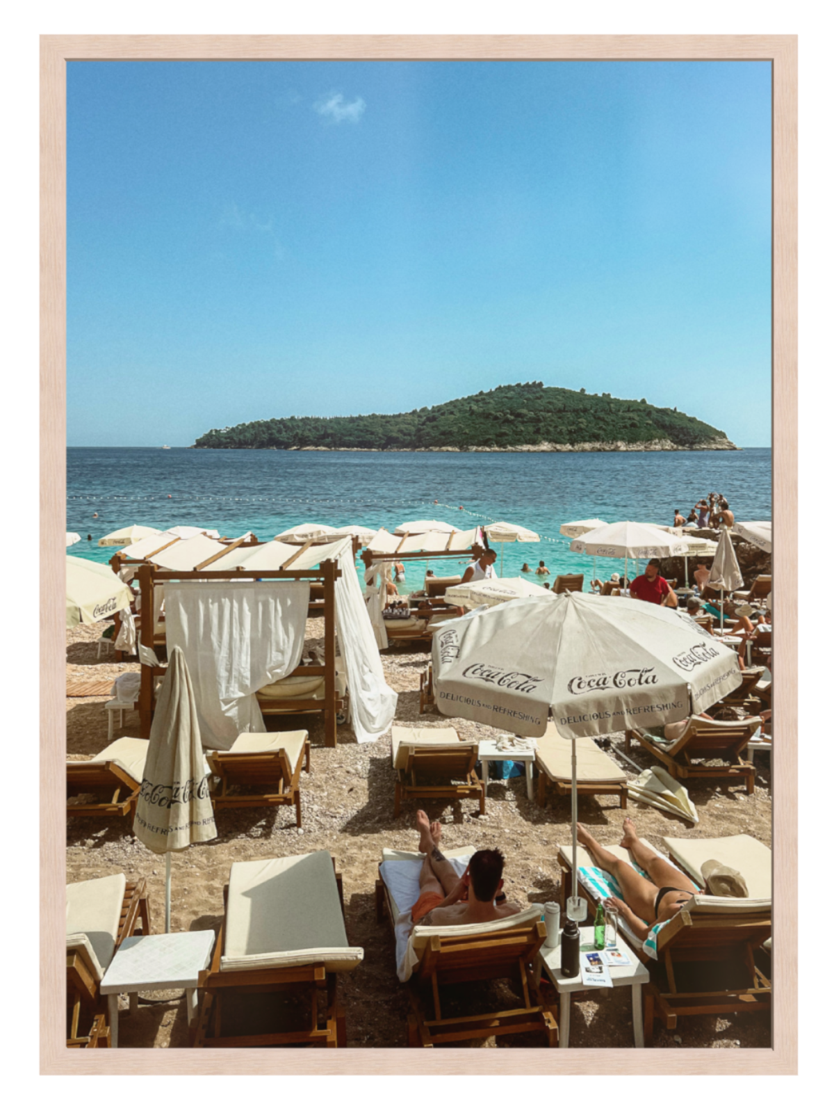 Beach scene with people, umbrellas, and a distant island under a clear blue sky.