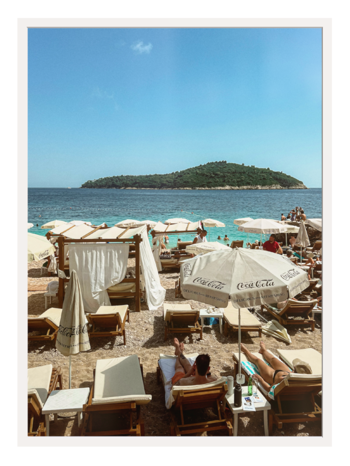 Beach scene with lounge chairs, umbrellas, and a distant island under a clear blue sky.