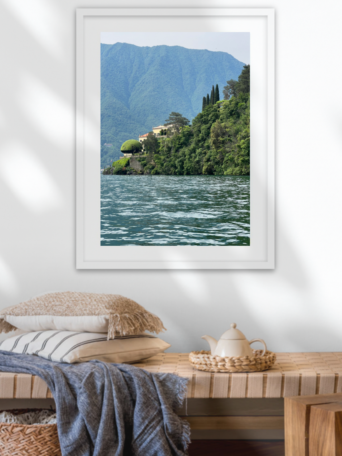 Framed landscape print of a mountainous lake scene above a wooden table with pillows and a teapot.