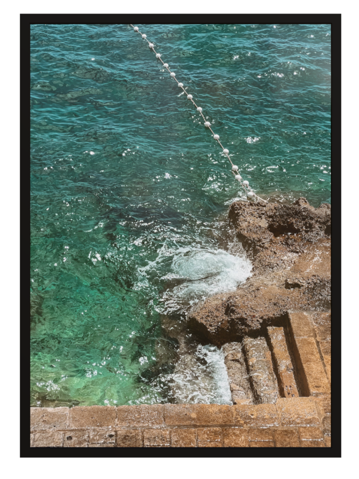 Turquoise ocean water crashing against a rocky shore with a wooden walkway. black frame