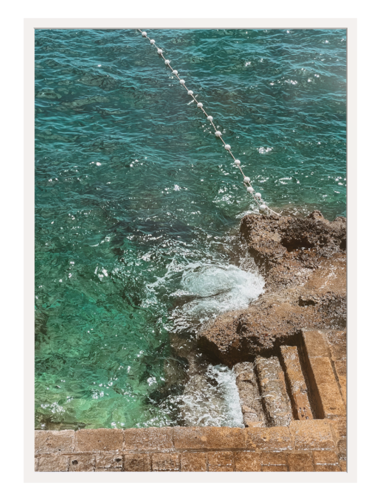 Turquoise ocean water crashing against a rocky shoreline with a rope barrier. white frame
