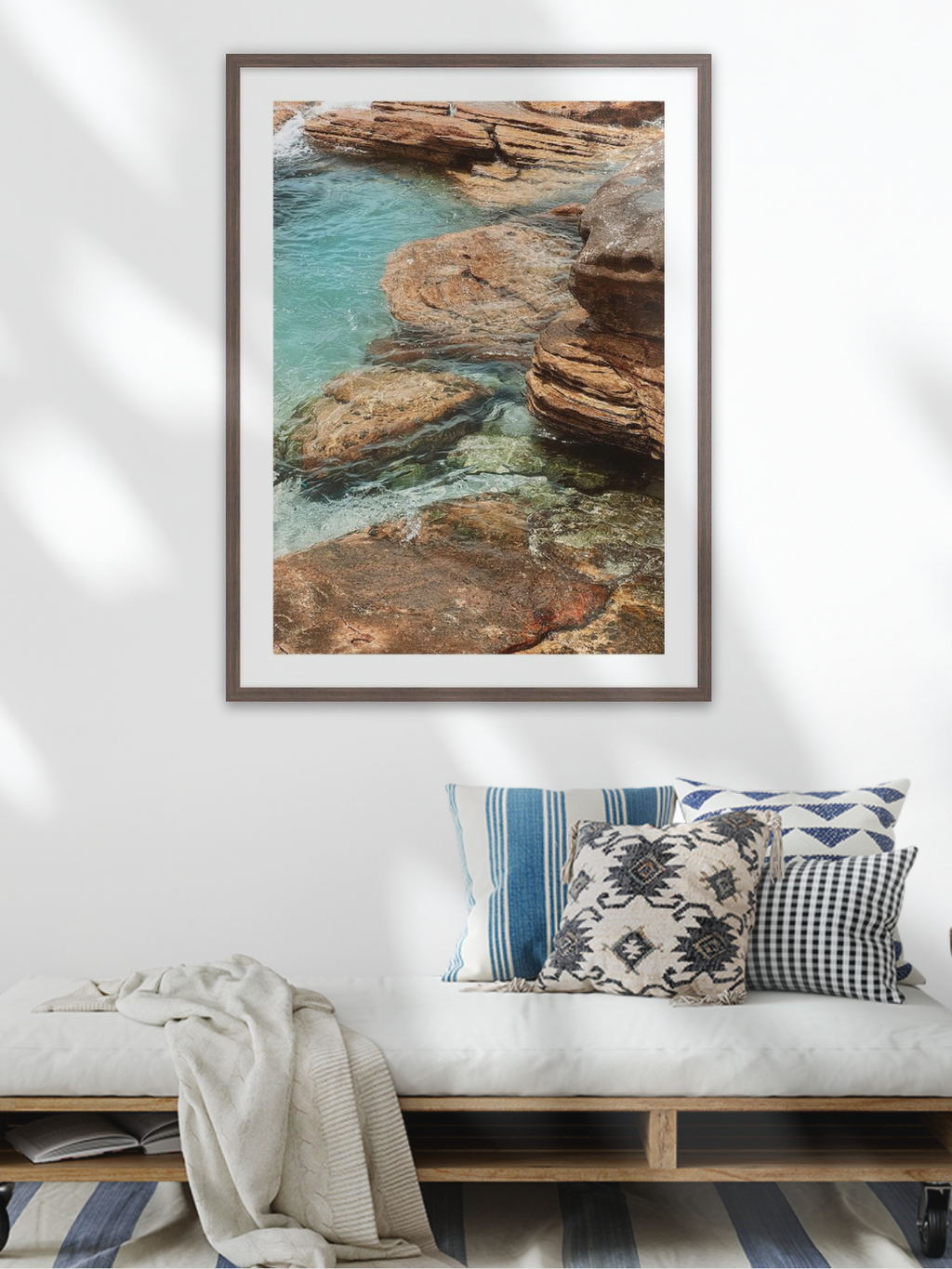 Framed coastal artwork above a bench with decorative pillows in a living room setting.